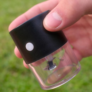 Puede incluir: Un pequeño picador de alimentos eléctrico portátil con una carcasa superior negra mate, un botón de encendido circular blanco y una base de recipiente de plástico transparente y cilíndrico. Se ve una cuchilla de metal dentro.