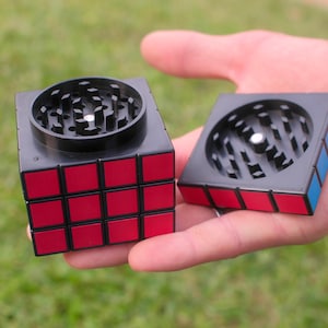 Puede incluir: Un molinillo de hierbas de dos piezas con forma de cubo y un patrón de cuadrícula rojo y negro en el exterior. La sección superior está separada, revelando un interior negro con dientes afilados en forma de pirámide para moler.