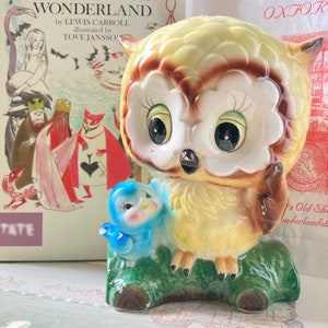 May include: A ceramic owl figurine with a bluebird perched on its side. The owl has large, black eyes, and is primarily yellow and brown. The figurine sits on a green log base. The background includes a book cover with the title "WONDERLAND".