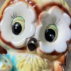 May include: A ceramic owl figurine with large, glossy black eyes and a detailed, colorful design. The owl has a yellow and brown body with white accents around the eyes. The figurine has a small, blue bird on its chest.