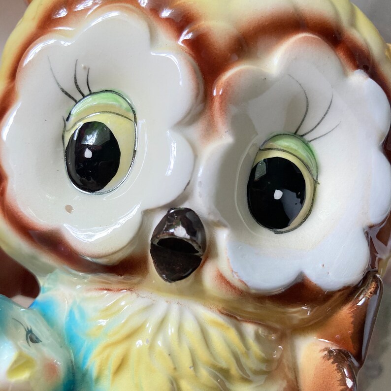 May include: A ceramic owl figurine with large, glossy black eyes and a detailed, colorful design. The owl has a yellow and brown body with white accents around the eyes. The figurine has a small, blue bird on its chest.