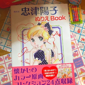 May include: A Japanese coloring book featuring a manga-style illustration of a young woman with blonde hair and a polka-dot dress. The cover includes Japanese text and the title "ぬりえ Book". A pack of colorful crayons is visible.
