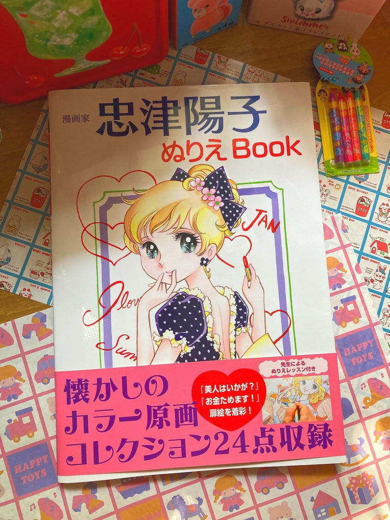 May include: A Japanese coloring book featuring a manga-style illustration of a young woman with blonde hair and a polka-dot dress. The cover includes Japanese text and the title "ぬりえ Book". A pack of colorful crayons is visible.