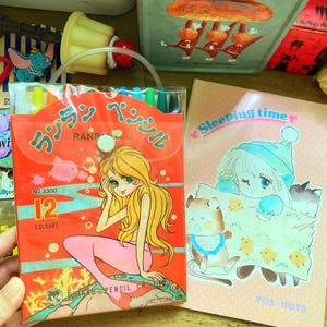 May include: A vintage pencil case with a mermaid illustration and the text "12 COLOURS". Next to it is a pink notebook with the text "Sleeping time" and an illustration of a girl in a hat with animals. The case contains colorful pens.