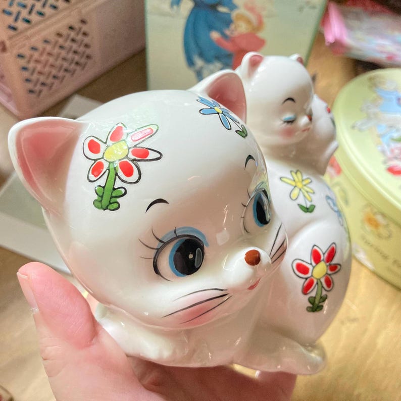May include: A white ceramic cat figurine with painted flowers and blue eyes. The cat is sitting with its head tilted to the side and its paws tucked under its body.