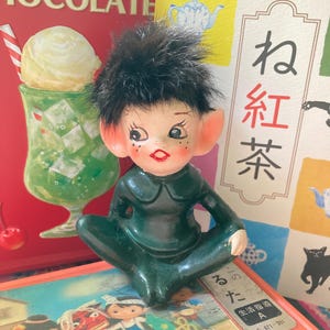 Könnte beinhalten: Eine kleine Figur in einem dunkelgrünen Outfit mit spitzen Ohren und schwarzem Haar. Das bemalte Gesicht zeigt große Augen, Sommersprossen und rote Lippen. Die Figur sitzt im Schneidersitz mit den Armen auf den Knien und hat einen neutralen Ausdruck.