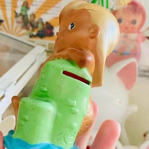 May include: A vintage mermaid figurine, with light brown skin, blonde hair, and a green coin bank. The mermaid sits on a blue base, with a red coin slot. This collectible figurine is a retro item.