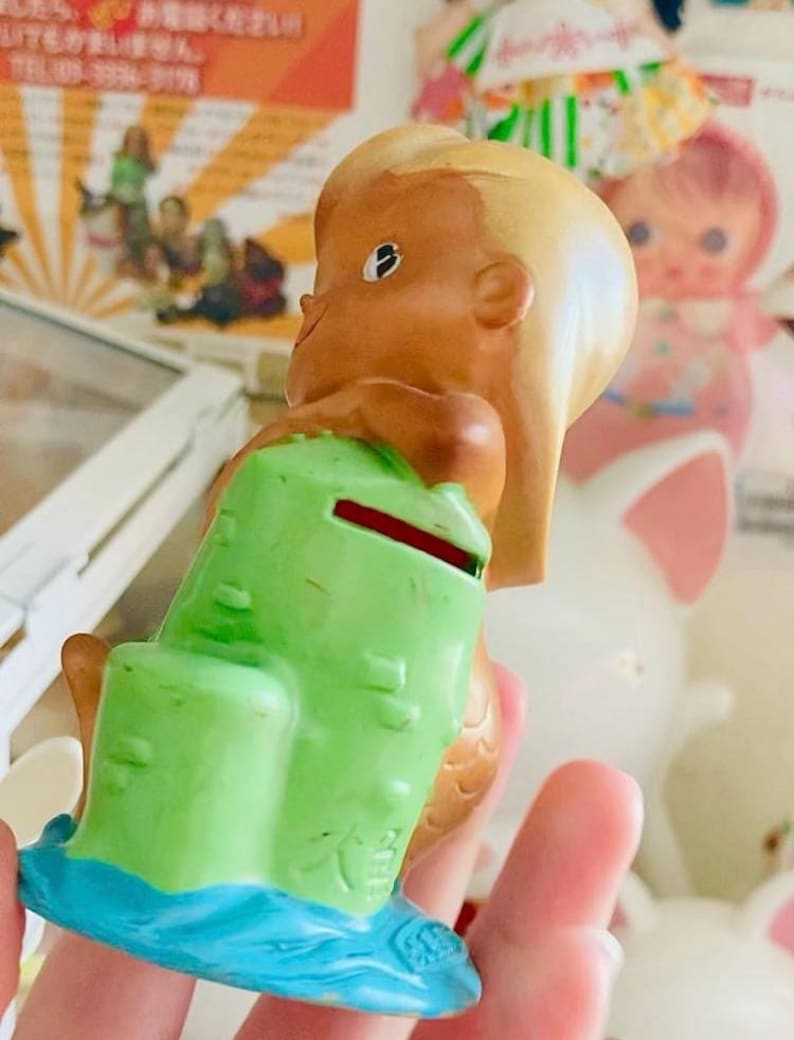 May include: A vintage mermaid figurine, with light brown skin, blonde hair, and a green coin bank. The mermaid sits on a blue base, with a red coin slot. This collectible figurine is a retro item.