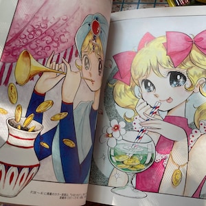 May include: A colorful illustration from a book featuring two anime-style characters. One character is playing a golden horn, with coins falling into a decorative vase. The other character is drinking from a glass with coins inside.