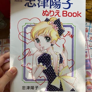 May include: A coloring book with a manga-style illustration on the cover. The image features a blonde-haired girl in a polka-dot dress, holding a lipstick. The book's title is in Japanese, with the word "Book" in English.