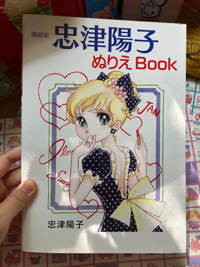 May include: A coloring book with a manga-style illustration on the cover. The image features a blonde-haired girl in a polka-dot dress, holding a lipstick. The book's title is in Japanese, with the word "Book" in English.