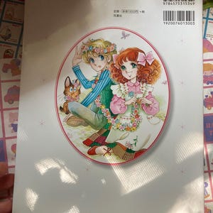 May include: Back cover of a Japanese manga book featuring a colorful illustration of two young people with floral garlands and a cat. The book has a white background with text and a barcode.