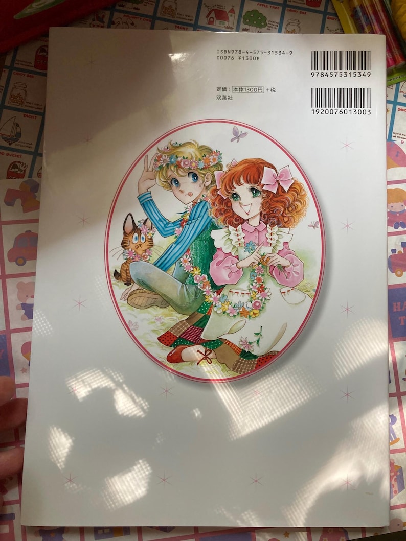 May include: Back cover of a Japanese manga book featuring a colorful illustration of two young people with floral garlands and a cat. The book has a white background with text and a barcode.