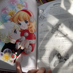 May include: A colorful illustration from a coloring book features two anime-style characters embracing. One has blonde hair and wears a black sweater, while the other has red hair and a red outfit. The background includes flowers and text.