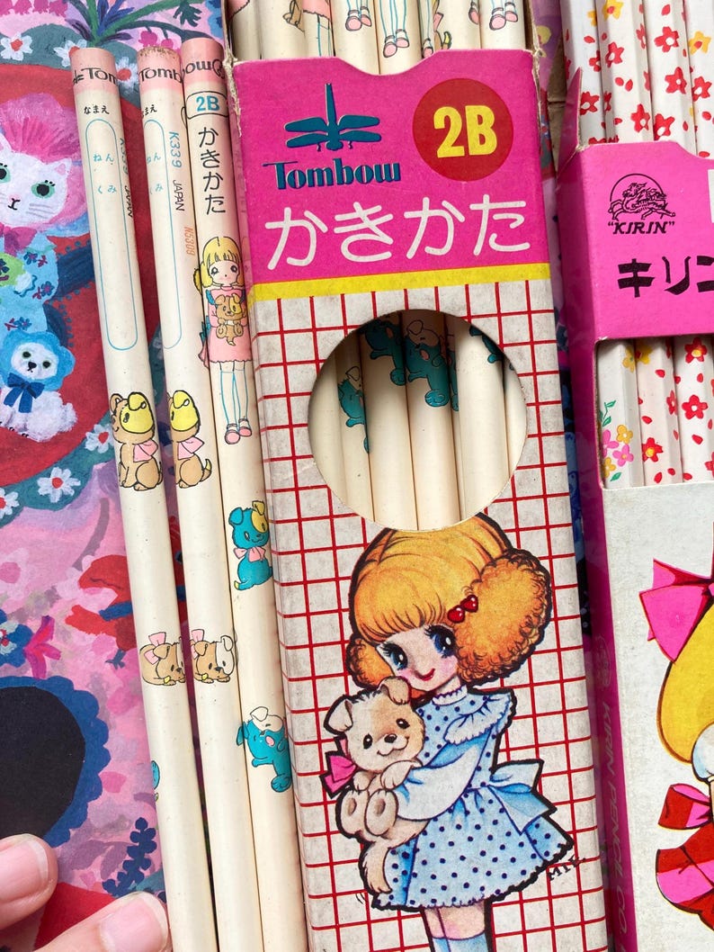 Puede incluir: Una colecci&oacute;n de l&aacute;pices y cajas de l&aacute;pices vintage. Los l&aacute;pices son blancos con ilustraciones de dibujos animados de cachorros y ni&ntilde;as. Las cajas de l&aacute;pices son rosas y presentan texto japon&eacute;s e ilustraciones de una ni&ntilde;a sosteniendo un cachorro.
