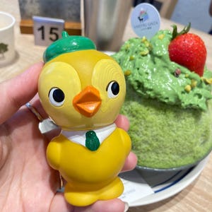 May include: A yellow chick figurine wearing a green hat and tie, with an orange beak and black eyes. The figurine is held in front of a green dessert topped with a strawberry. A metal cup and a table number are in the background.