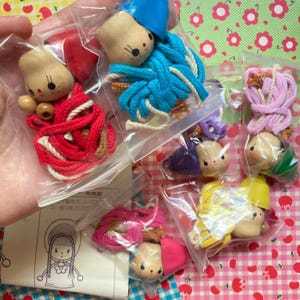 May include: A collection of small, handmade doll heads with various colored yarn and accessories, packaged in clear plastic bags. The dolls have painted faces and are adorned with red, blue, pink, and yellow yarn. The background features a floral pattern.