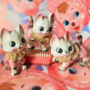 May include: Three ceramic cat figurines with pink and white striped bodies, large blue eyes, and tan lace collars. The cats are posed on a pink and red patterned background.