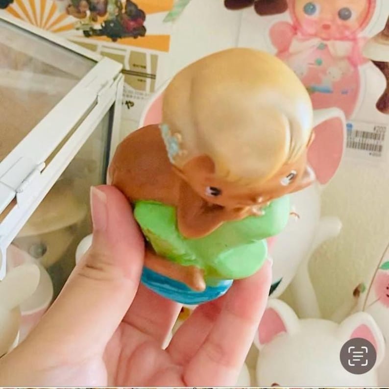 May include: A vintage-style figurine of a baby mermaid with blonde hair, brown body, and green and blue accents. The figurine is held in a hand, with other toys in the background.