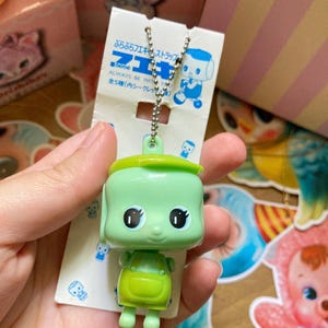 May include: A green plastic keychain featuring a cartoon character with large eyes and a smiling face. The character wears a green outfit and a matching hat. The keychain is attached to a silver chain and a white tag with Japanese text.