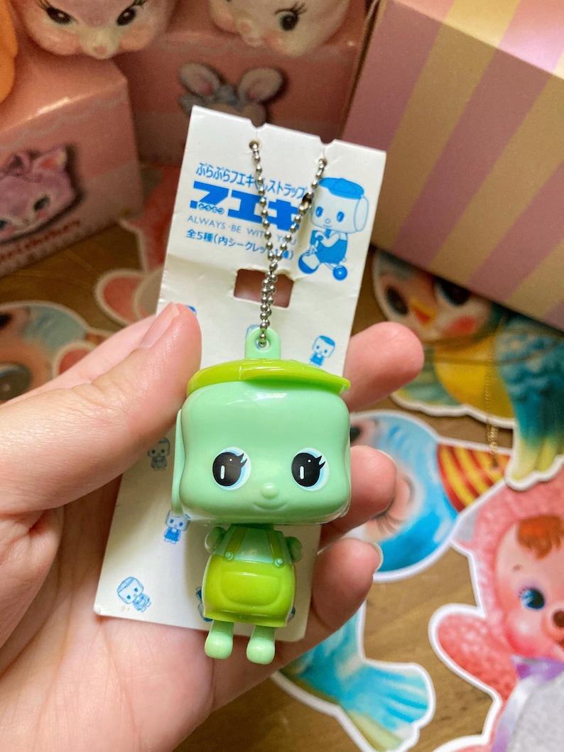 May include: A green plastic keychain featuring a cartoon character with large eyes and a smiling face. The character wears a green outfit and a matching hat. The keychain is attached to a silver chain and a white tag with Japanese text.