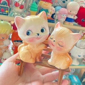 May include: Two vintage ceramic cat figurines, one sitting and one leaning, on a small wooden bench. The cats have peach-colored fur, pink cheeks, and blue eyes. The figurines are part of a collection of vintage decor.