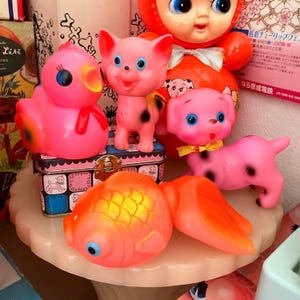 May include: A selection of vintage pink rubber toys, including a duck, pig, dog, and goldfish. The toys have blue eyes and black spots. A red-orange doll is in the background. The toys are on a white surface.