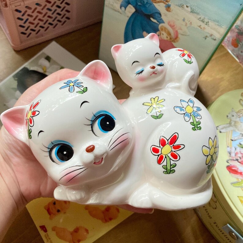 May include: A white ceramic cat figurine with a smaller white cat figurine on its back. Both cats have painted blue eyes, pink noses, and black whiskers. The larger cat has colourful flowers painted on its body.