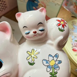 May include: A white ceramic money box shaped like two kittens cuddling. The kittens have painted blue eyes, pink noses, and black eyelashes. The money box is decorated with colourful flowers.