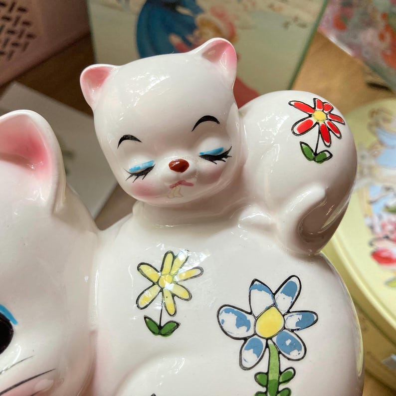 May include: A white ceramic money box shaped like two kittens cuddling. The kittens have painted blue eyes, pink noses, and black eyelashes. The money box is decorated with colourful flowers.