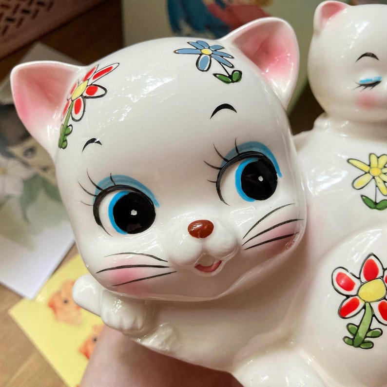 May include: A white ceramic cat figurine with blue eyes, pink cheeks, and a red, yellow, and blue flower design. The cat is sitting with its head tilted and has a playful expression.
