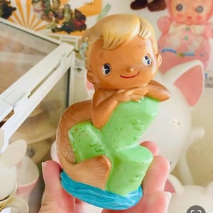 Puede incluir: Una figura de sirena de estilo vintage con una cara sonriente, cabello rubio y un cuerpo marrón. La sirena está posada sobre una roca verde con una base azul. La figura está hecha de un material brillante.