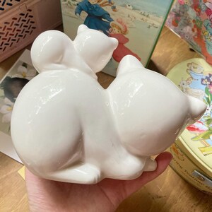 May include: A white ceramic figurine of two cats, one sitting on top of the other. The cats are facing forward and have a glossy finish.
