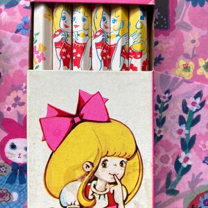 Puede incluir: Juego de papeler&iacute;a vintage con una caja que presenta una ni&ntilde;a de dibujos animados con un lazo rosa y l&aacute;pices con un dise&ntilde;o similar. Los l&aacute;pices tienen una ilustraci&oacute;n repetida de una ni&ntilde;a con un vestido rojo.