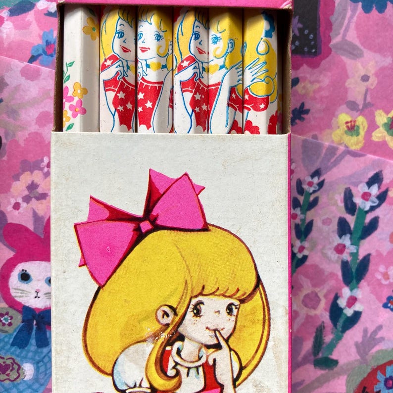 Puede incluir: Juego de papeler&iacute;a vintage con una caja que presenta una ni&ntilde;a de dibujos animados con un lazo rosa y l&aacute;pices con un dise&ntilde;o similar. Los l&aacute;pices tienen una ilustraci&oacute;n repetida de una ni&ntilde;a con un vestido rojo.