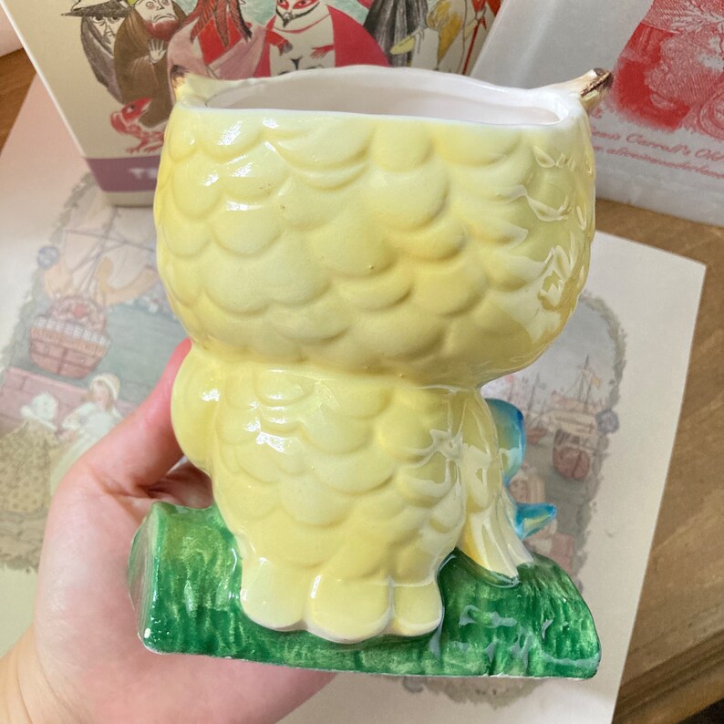 May include: A yellow ceramic owl-shaped planter with textured feather details. The owl is perched on a green base with blue accents. The planter has a white interior and gold-colored accents on the ears. A whimsical decorative item.