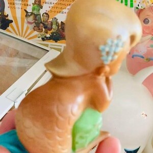 May include: A vintage mermaid figurine with a tan head and brown body, decorated with a light blue flower on the side of the head. The mermaid's tail is green and blue. The figurine is held in a hand, with other toys in the background.