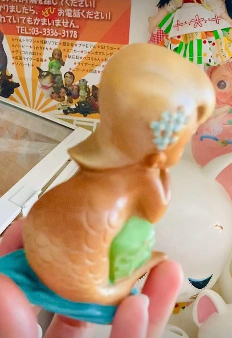 May include: A vintage mermaid figurine with a tan head and brown body, decorated with a light blue flower on the side of the head. The mermaid's tail is green and blue. The figurine is held in a hand, with other toys in the background.