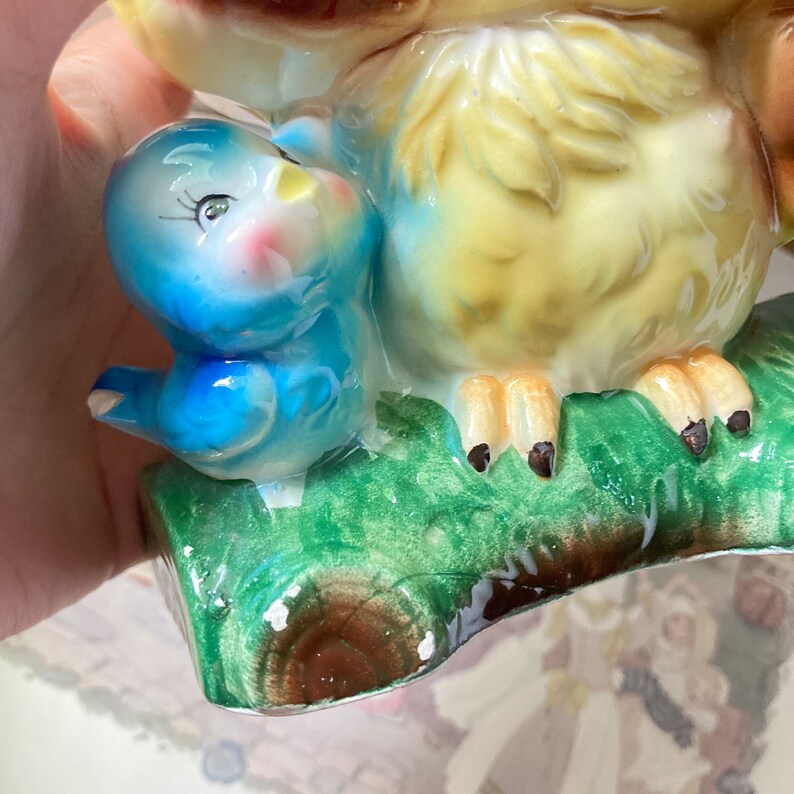 May include: A vintage ceramic figurine of a large owl with a small bluebird perched beside it. The owl has yellow and brown feathers, while the bluebird has a blue body and pink cheeks. The figurine is set on a green branch with brown accents.
