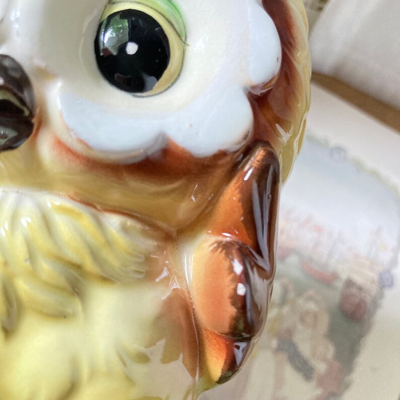 May include: Close-up of a ceramic owl figurine. The owl has large, glossy black eyes with green accents, and a white and brown feathered design. The owl's beak and ear tufts are brown, and the body is yellow.
