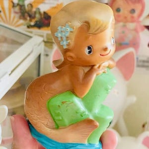 May include: A vintage mermaid figurine with blonde hair and blue flower accents. The mermaid has a brown tail and is sitting on a green object, with a blue base. The figurine is held in a hand.