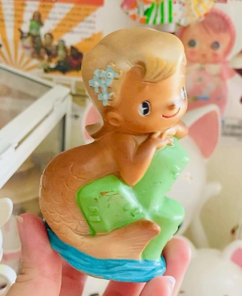 May include: A vintage mermaid figurine with blonde hair and blue flower accents. The mermaid has a brown tail and is sitting on a green object, with a blue base. The figurine is held in a hand.