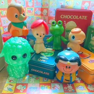 May include: A selection of brightly coloured vintage plastic toys, including a green sheep, a frog, a duck, and a figure with red hair. A red tin with "CHOCOLATE" is visible in the background. The toys are arranged on a patterned surface.