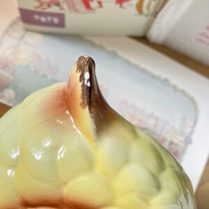 May include: Close-up of a ceramic object with a rounded, textured body in shades of yellow and cream. The top portion is a pointed, brown-tipped shape. The object appears to be part of a larger decorative piece, with a blurred background.