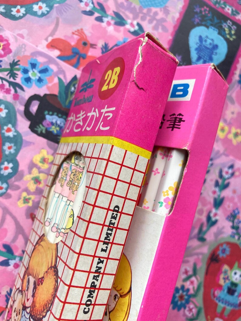 Puede incluir: Dos cajas de l&aacute;pices, una rosa brillante y otra magenta, con texto e ilustraciones japonesas. Las cajas est&aacute;n sobre un fondo floral rosa. Una caja est&aacute; etiquetada como "2B" y la otra como "B". Los l&aacute;pices del interior son visibles.