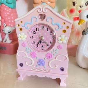 May include: A pink, decorative clock shaped like a house with a white face and black hands. The clock has a floral design with pink, purple, and white flowers. The clock face has the word "Swimmer" written on it.