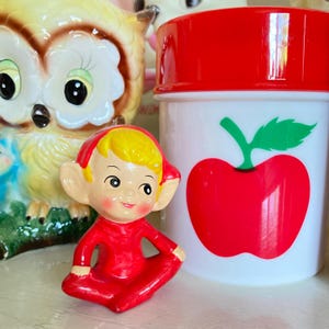Puede incluir: Una figura roja de un elfo sentado con cabello amarillo y orejas grandes. El elfo lleva un atuendo rojo y una gorra roja. Detrás del elfo hay un recipiente blanco con tapa roja y un gráfico de manzana roja. Una figura de búho de cerámica está en el fondo.