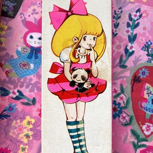 Puede incluir: Caja de l&aacute;pices vintage con una ilustraci&oacute;n de una ni&ntilde;a de dibujos animados. La ni&ntilde;a tiene el pelo rubio, un lazo rosa y un vestido a rayas rojas y rosas, sosteniendo un panda. El fondo es rosa con motivos florales y de conejos.