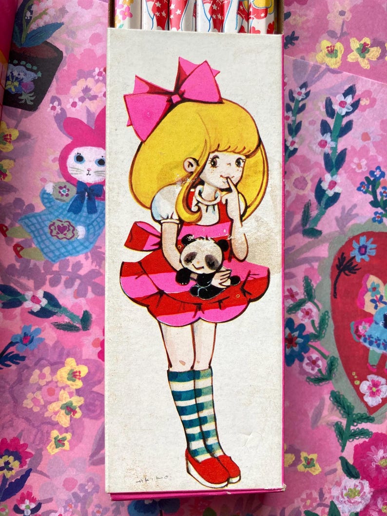 Puede incluir: Caja de l&aacute;pices vintage con una ilustraci&oacute;n de una ni&ntilde;a de dibujos animados. La ni&ntilde;a tiene el pelo rubio, un lazo rosa y un vestido a rayas rojas y rosas, sosteniendo un panda. El fondo es rosa con motivos florales y de conejos.