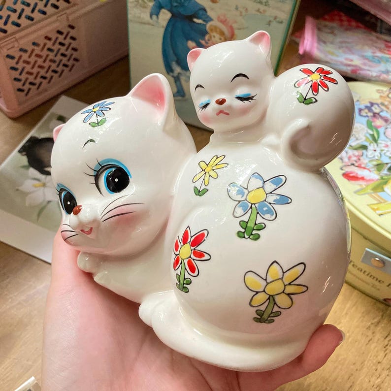 May include: White ceramic cat figurine with a smaller cat figurine on its back. Both cats have blue eyes and pink noses. The larger cat is decorated with colourful flowers.